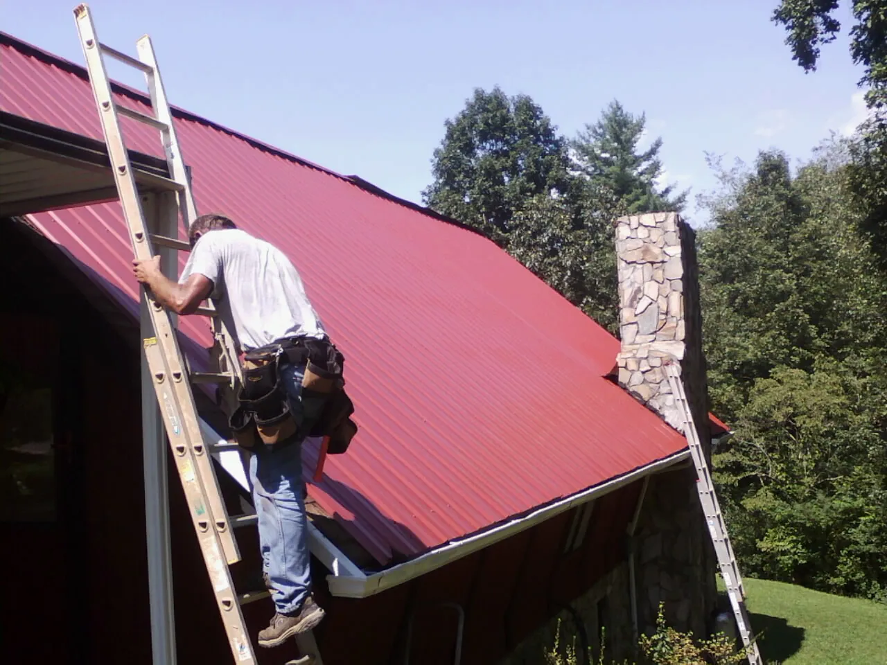 Roofer on a ladder beside a red metal roof and stone chimney — Soffit & Fascia Repair by Roofing Brothers in Neversink